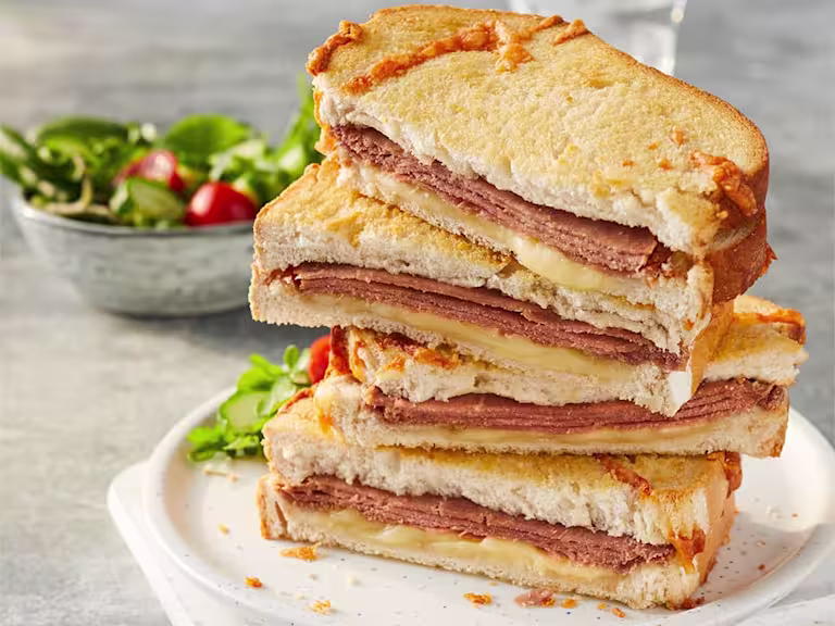 Four Quorn Vegetarian Ham And Cheese Toastie sandwich halves stacked on top of each other to show the filling with a side salad in the back.
