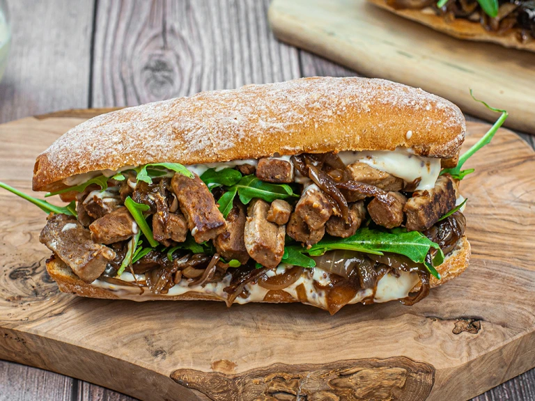 Steak strips and caramelised onions sandwich served on a wooden slab.