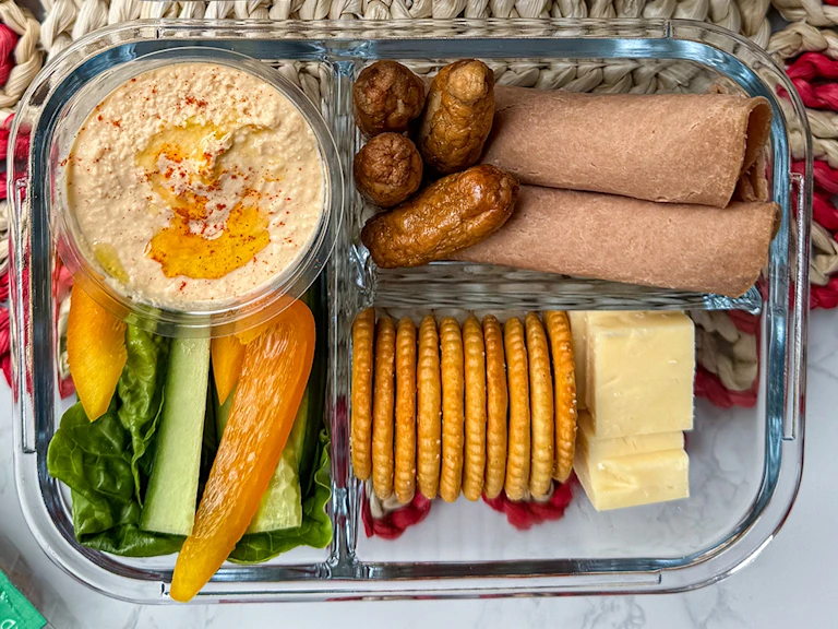 Vegetarian adult lunchbox with Quorn cocktail sausages, wholemeal wraps, cheddar cheese cubes, crackers, hummus, and sliced vegetables
