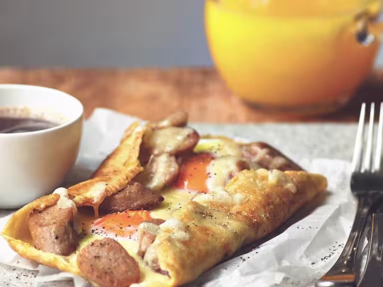 A galette filled with Quorn Sausage, cheese, and two eggs on a piece of paper with a cup of coffee and cutlery on either side.