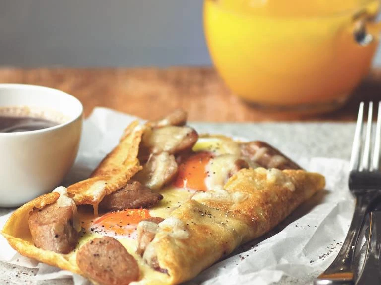 A galette filled with Quorn Sausage, cheese, and two eggs on a piece of paper with a cup of coffee and cutlery on either side.