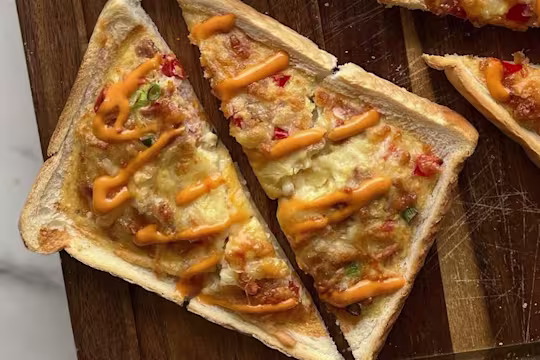 Baked toast topped with an egg, cheese, vegetarian ham and egg mixture drizzled with siracha mayo on a wooden board.