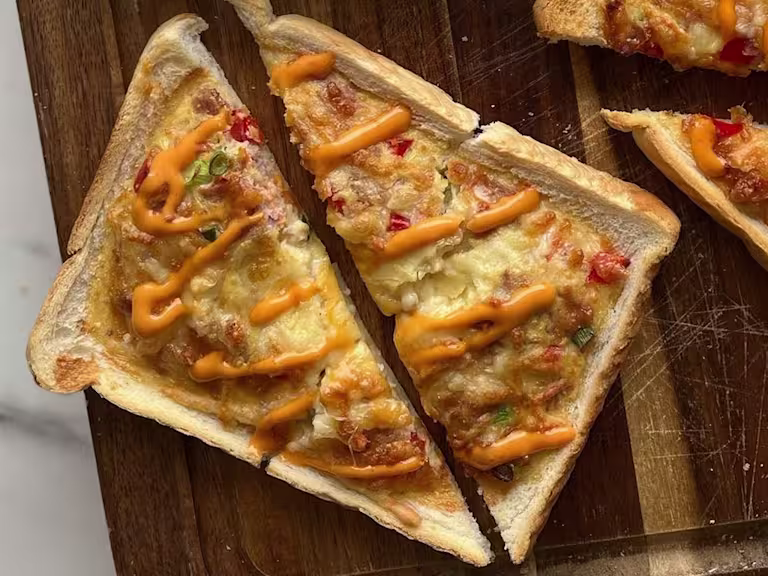 Baked toast topped with an egg, cheese, vegetarian ham and egg mixture drizzled with siracha mayo on a wooden board.