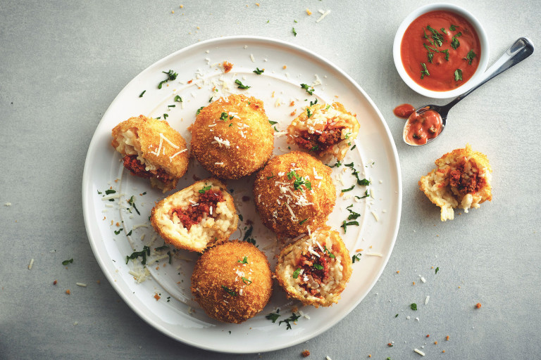 Five arancini on a white plate, two of white have been halved to show they're filled with Quorn Grounds, with a small bowl of tomato sauce on the side.