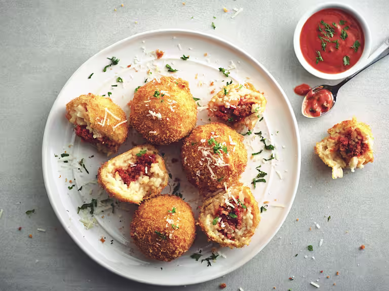 Five arancini on a white plate, two of white have been halved to show they're filled with Quorn Grounds, with a small bowl of tomato sauce on the side.