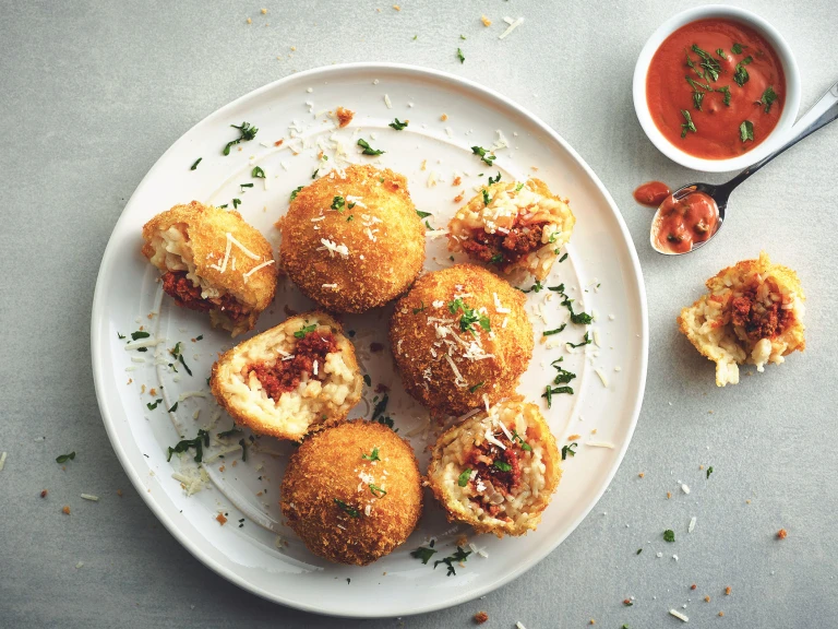 Five arancini on a white plate, two of white have been halved to show they're filled with Quorn Grounds, with a small bowl of tomato sauce on the side.