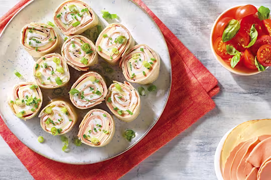 Eleven slices of a vegan ham and cheese roll made with Quorn Vegan Smoky Ham Free Slices, Violife Smoky Cheddar Flavour Slices, Violife Creamy, and chives topped with sliced spring onions.