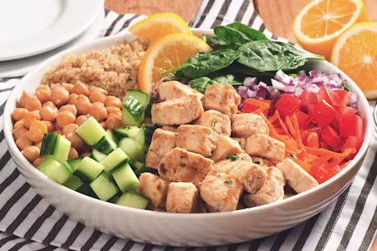 A white bowl with quinoa, spinach, diced red onion, shredded carrot, Quorn Pieces, diced cucumber, and chickpeas arranged in sections around the bowl and topped with two slices of lemon.