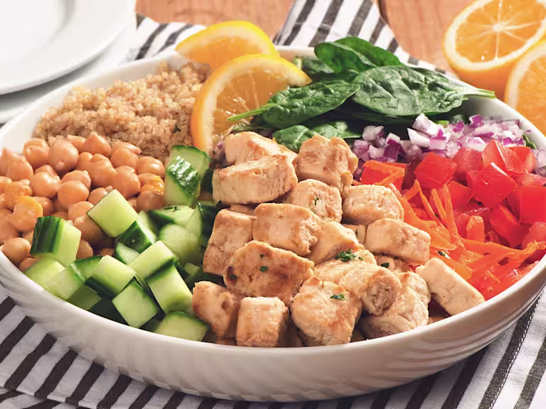 A white bowl with quinoa, spinach, diced red onion, shredded carrot, Quorn Pieces, diced cucumber, and chickpeas arranged in sections around the bowl and topped with two slices of lemon.
