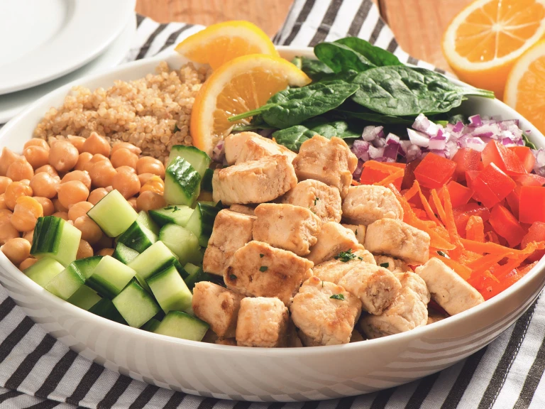 A white bowl with quinoa, spinach, diced red onion, shredded carrot, Quorn Pieces, diced cucumber, and chickpeas arranged in sections around the bowl and topped with two slices of lemon.