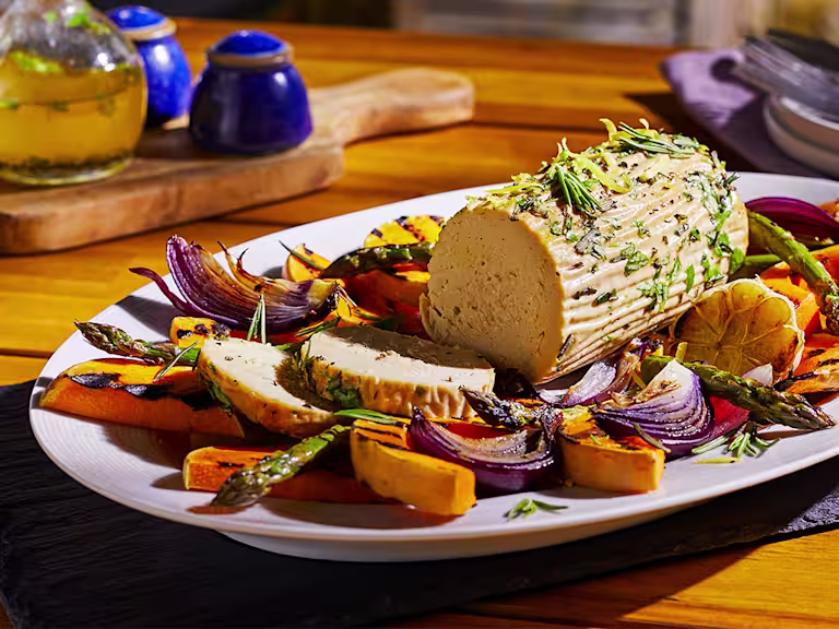 A summer vegetarian roast made with Quorn Meatless Roast topped with rosemary and lemon zest in the centre of a platter surrounded by roasted vegetables.