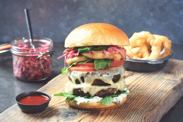 A double Quorn burger topped with cheese, arugula, tomatoes, jalapenos, pink slaw, and onion rings.