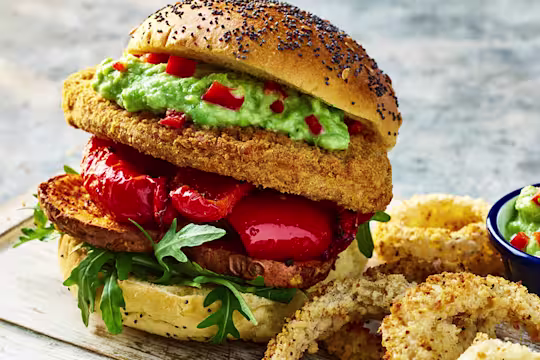 Two Quorn Crunchy Filet Cajun Burgers with onion rings on a board with a sweat pea guacamole in a bowl on the side