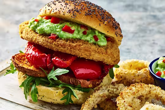 Two Quorn Crunchy Filet Cajun Burgers with onion rings on a board with a sweat pea guacamole in a bowl on the side