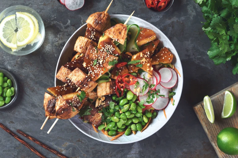 A bowl filled a salad of quinoa, lettuce, red cabbage, red pepper, carrot, spring onion, edamame, cucumber, and radish, topped with two skewers of grilled Quorn Fillet pieces and pineapple and coriander, chilli, sesame seeds, peanuts, and wedges of lime.