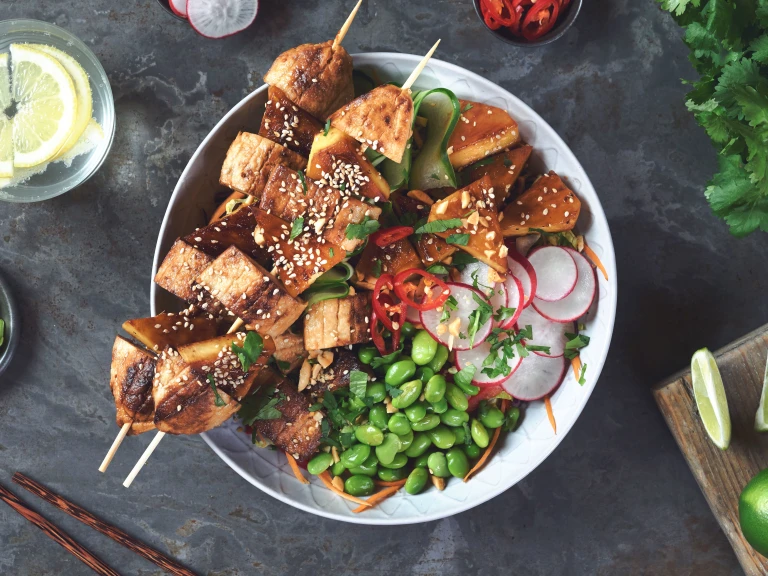 A bowl filled a salad of quinoa, lettuce, red cabbage, red pepper, carrot, spring onion, edamame, cucumber, and radish, topped with two skewers of grilled Quorn Fillet pieces and pineapple and coriander, chilli, sesame seeds, peanuts, and wedges of lime.