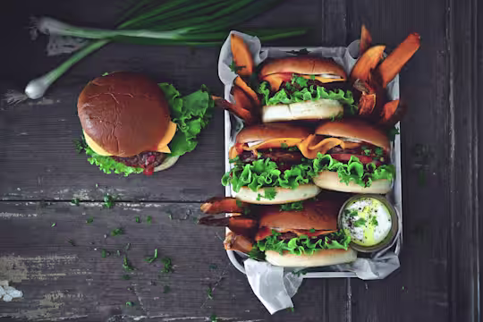 4 Quorn Quarter Pounders in buns served on a tray with sweet potato fries and a pot of mayonnaise with one burger on the table