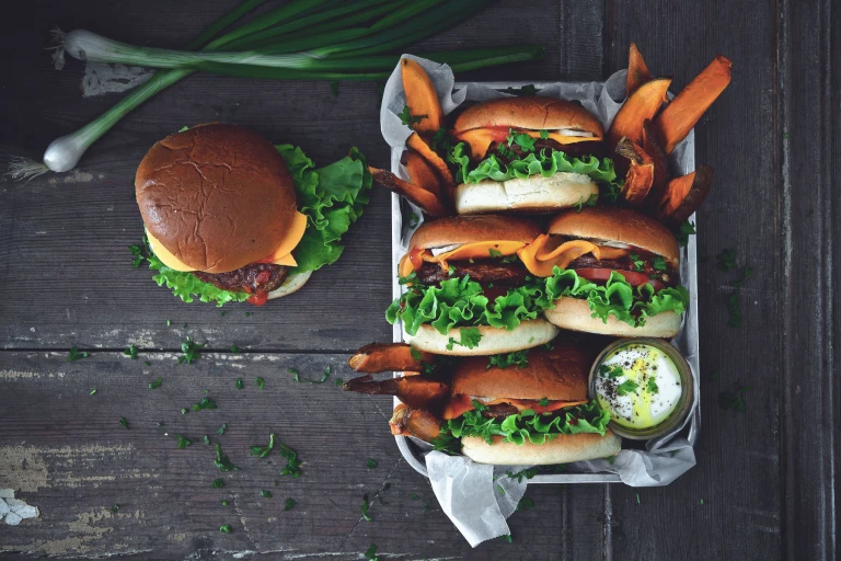 4 Quorn Classic Beef Style Burgers in buns served on a tray with sweet potato fries and a pot of mayonnaise with one burger on the table