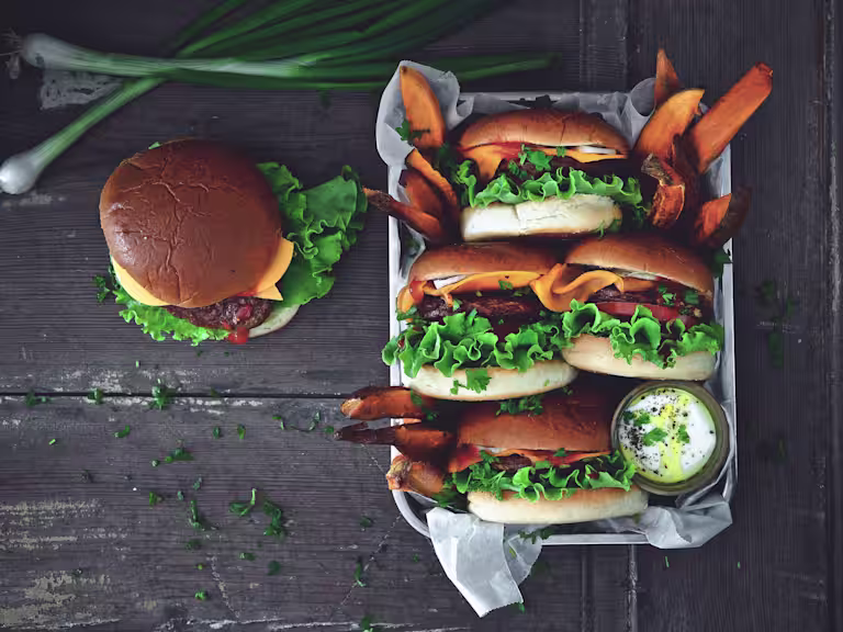 4 Quorn Quarter Pounders in buns served on a tray with sweet potato fries and a pot of mayonnaise with one burger on the table
