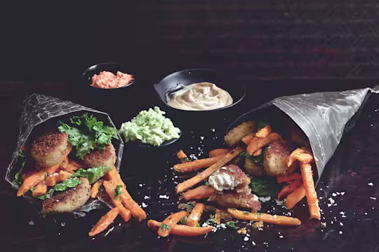 Two paper cones filled with Quorn Southern Fried Bites and sweet potato fries with three dips in the background.