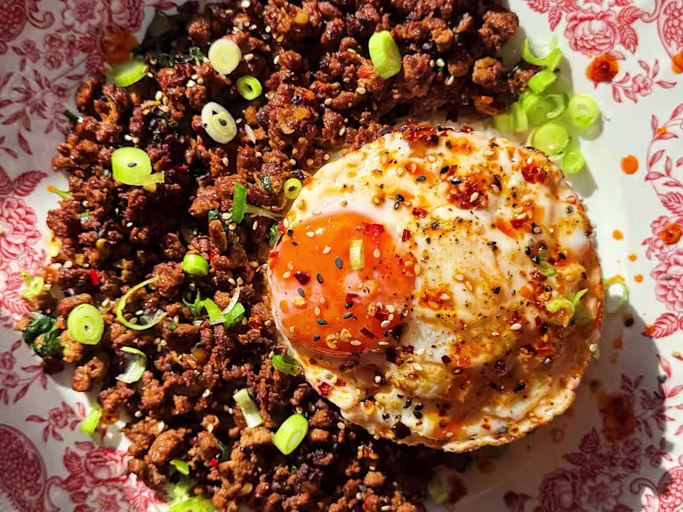 A bowl of Vegetarian Pad Kra Pao with Quorn Mince and garlic cloves, shallots, ginger, basil, sesame seeds and a fried egg on top.