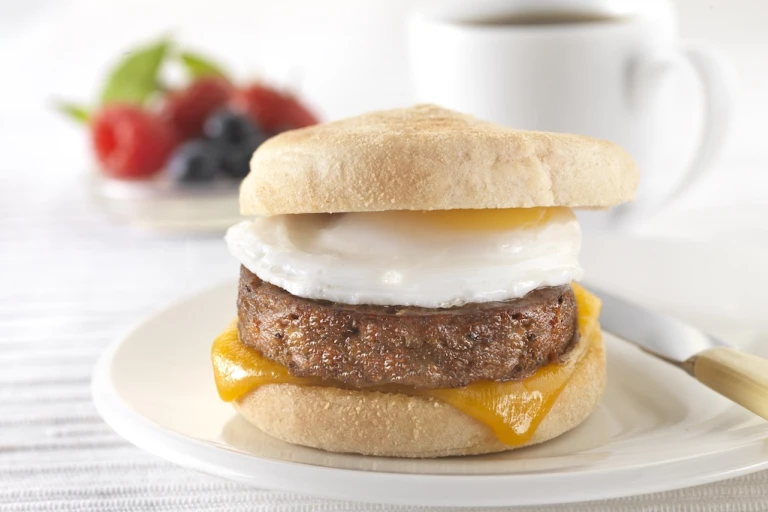 A muffin filled with a Quorn Sausage Patty, a slice of melted cheese, and a fried egg on a white plate.