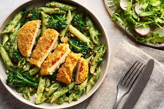 A bowl of penne pasta tossed in vibrant green broccoli pesto with tender florets, topped with a sliced Quorn Lemon and Pepper Schnitzel and a sprinkle of pine nuts.