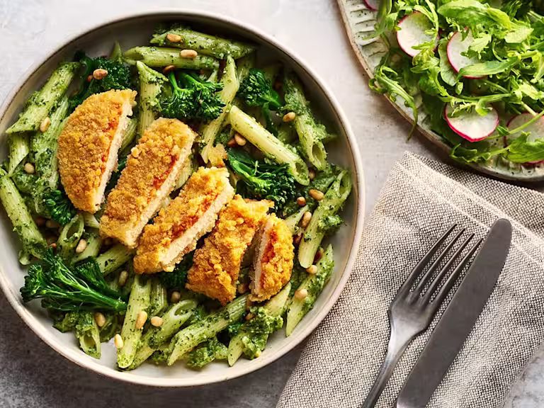 A bowl of penne pasta tossed in vibrant green broccoli pesto with tender florets, topped with a sliced Quorn Lemon and Pepper Schnitzel and a sprinkle of pine nuts.
