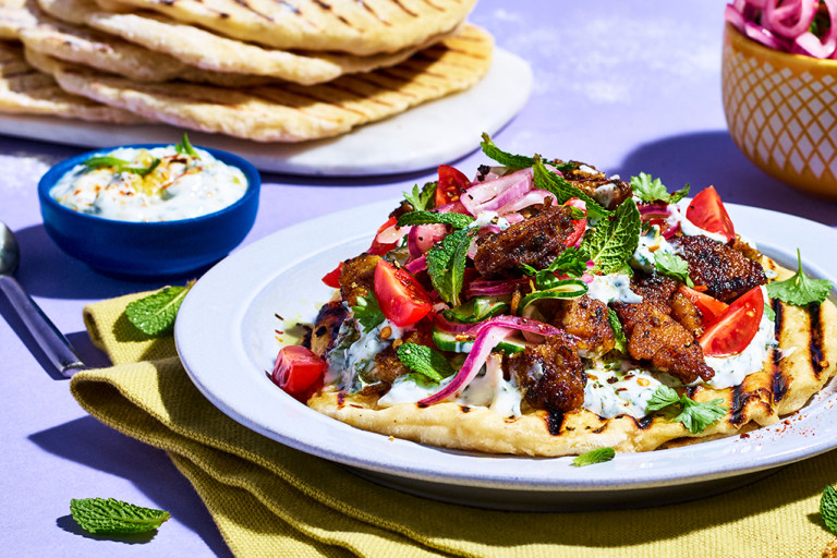 Flatbread topped with mint yogurt dressing, pickled red onions, cucumber, tomato, and Quorn Makes Amazing Peri Peri Strips on a yellow napkin on a lilac background.