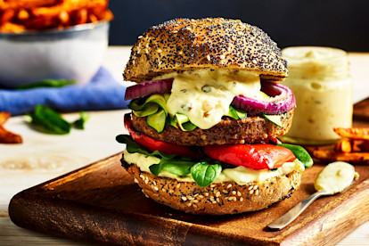 A Quorn Classic Beef Style Burger topped with red onion, red pepper, courgette, spinach, and burger sauce on a poppyseed bun on a board with fries on the side.