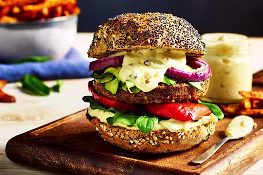 A Quorn Quarter Pounder topped with red onion, red pepper, courgette, spinach, and burger sauce on a poppyseed bun on a board with fries on the side.