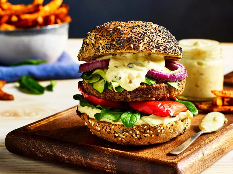 A Quorn Classic Beef Style Burger topped with red onion, red pepper, courgette, spinach, and burger sauce on a poppyseed bun on a board with fries on the side.