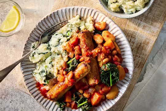 Quorn sausage cassoulet with chopped vegetables, beans, kale and mashed potato on a grey plate with a glass of water and lemon to the side.