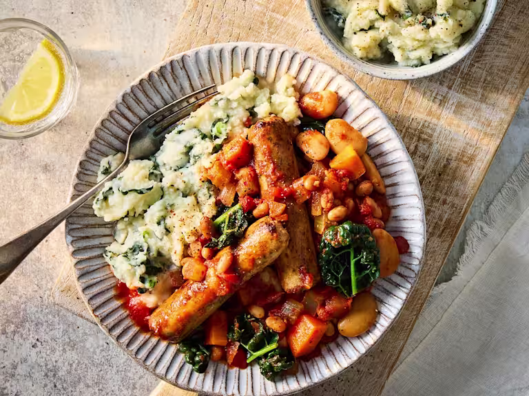 Quorn sausage cassoulet with chopped vegetables, beans, kale and mashed potato on a grey plate with a glass of water and lemon to the side.