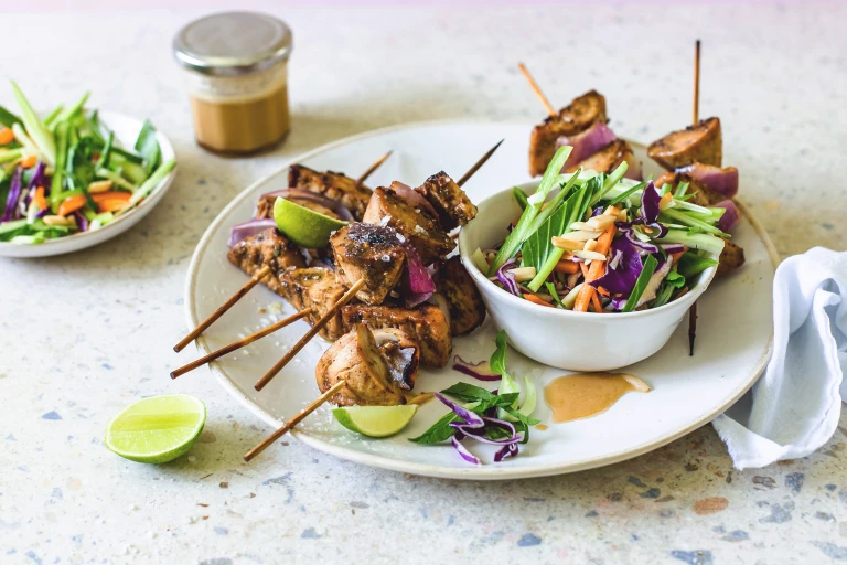 Quorn Pieces and red onion skewers piled high on a white plate with a bowl of red cabbage, carrot, cucumber, and bok choy slaw on the side with lime wedges for serving and dressing in the background.