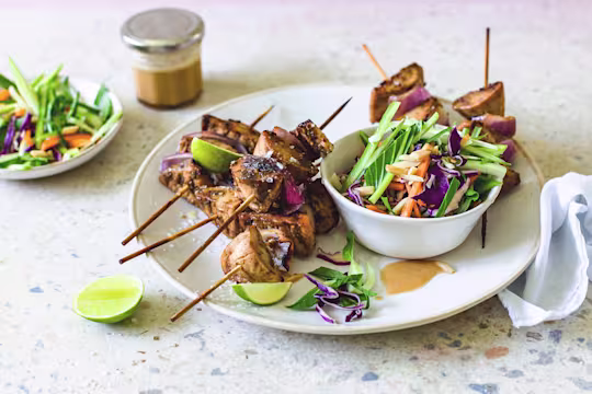 Quorn Fillets and red onion skewers piled high on a white plate with a bowl of red cabbage, carrot, cucumber, and bok choy slaw on the side with lime wedges for serving and dressing in the background.