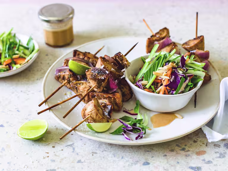 Quorn Fillets and red onion skewers piled high on a white plate with a bowl of red cabbage, carrot, cucumber, and bok choy slaw on the side with lime wedges for serving and dressing in the background.