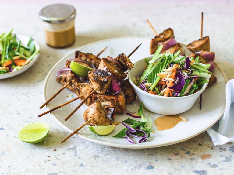 Quorn Pieces and red onion skewers piled high on a white plate with a bowl of red cabbage, carrot, cucumber, and bok choy slaw on the side with lime wedges for serving and dressing in the background.