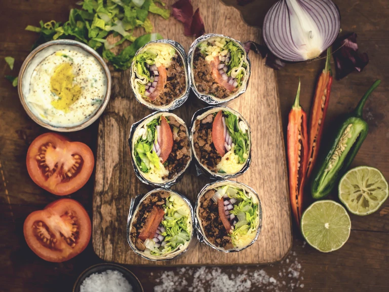 Three halved burritos filled with Quorn Grounds, red onion, tomato, and lettuce arranged on a plank with vegetables and a sauce placed around the board.