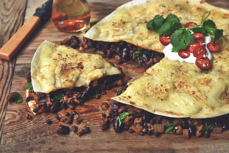 Quorn Mince, lentils, and black beans between two tortillas with cheese melted on top, garnished with sour cream, red chilli slices, and coriander on a cutting board.