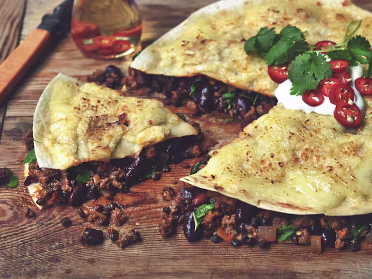 Quorn Mince, lentils, and black beans between two tortillas with cheese melted on top, garnished with sour cream, red chilli slices, and coriander on a cutting board.