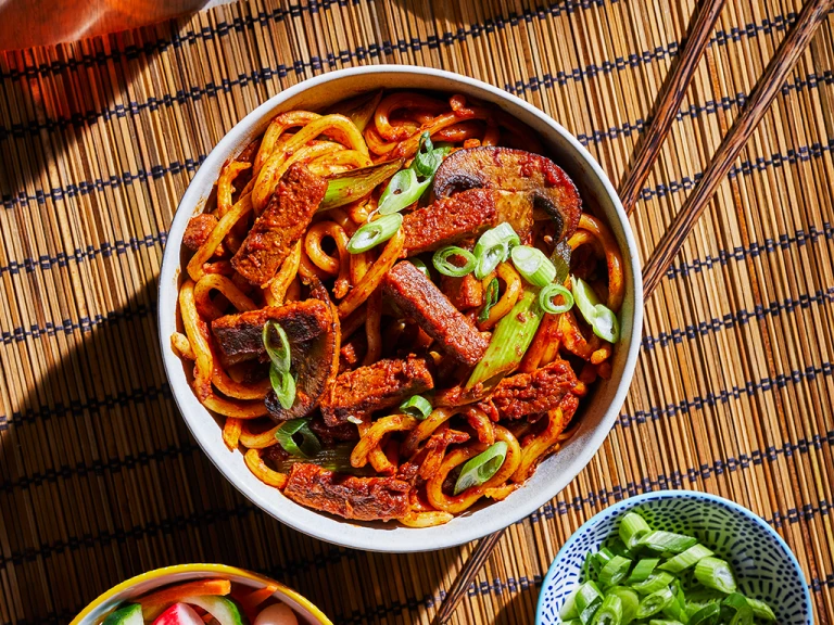 A bowl of vegetarian bulgogi noodles made with Quorn Steak Strips and udon noodles topped with scallions with a bowl of quick-pickled vegetables on the side.