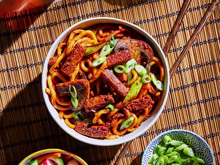A bowl of vegetarian bulgogi noodles made with Quorn Steak Strips and udon noodles topped with scallions with a bowl of quick-pickled vegetables on the side.