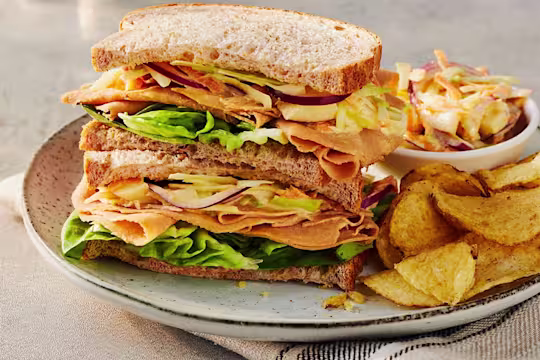 Two Quorn Finely Sliced Ham And Coleslaw Sandwich stacked on top of each other to display the filling served alongside a portion of coleslaw and crisps.