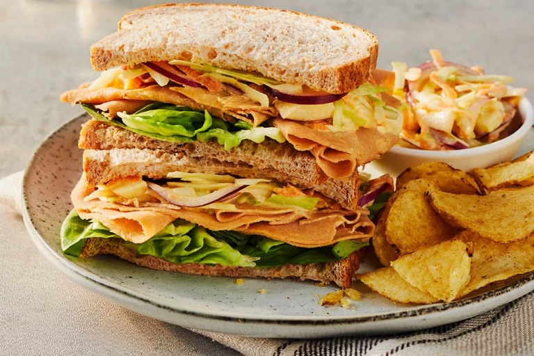 Two Quorn Finely Sliced Ham And Coleslaw Sandwich stacked on top of each other to display the filling served alongside a portion of coleslaw and crisps. 