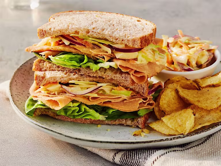 Two Quorn Finely Sliced Ham And Coleslaw Sandwich stacked on top of each other to display the filling served alongside a portion of coleslaw and crisps.
