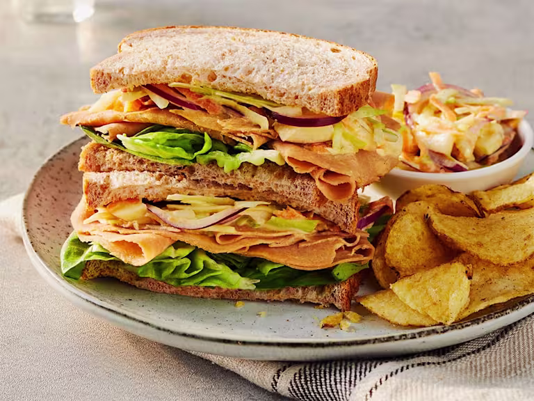 Two Quorn Finely Sliced Ham And Coleslaw Sandwich stacked on top of each other to display the filling served alongside a portion of coleslaw and crisps.