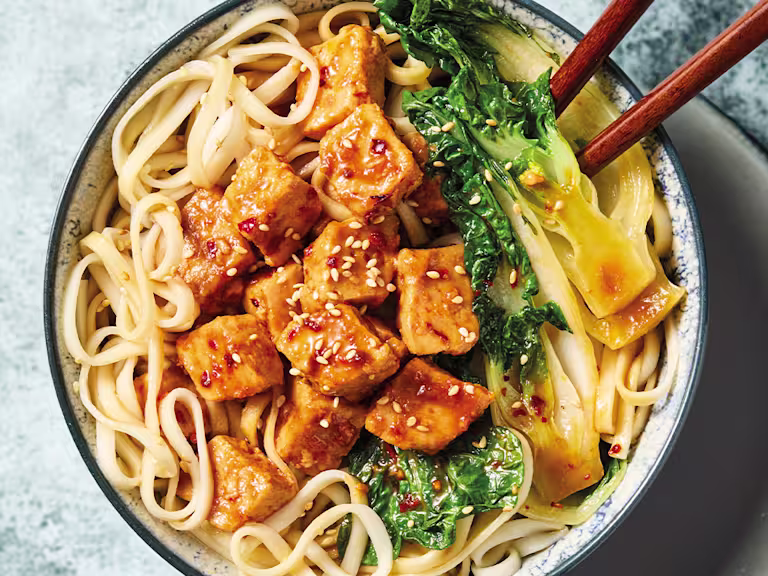 Yakisoba med Quorn Bitar och nudlar, grönsaker och ätpinnar, i en vit skål mot en blå bakgrund.