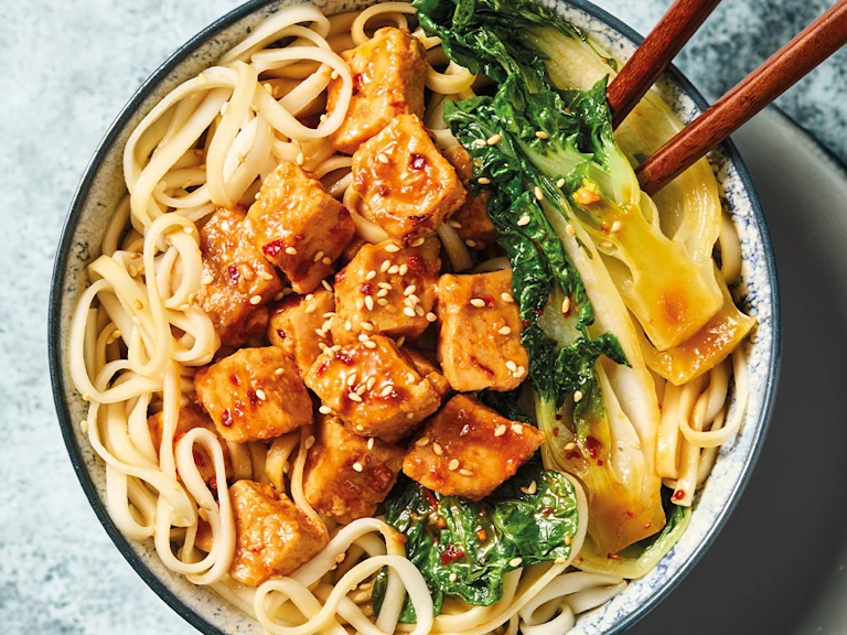 Quorn pieces Yakisoba with noodles, vegetables and chopsticks, in a white bowl on a blue background.