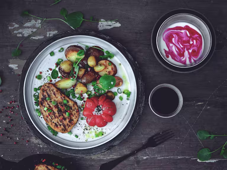 Quorn Vegetarian Peppered Steak, roast potatoes and tomato served on a plate dressed with salad leaves and a side of yoghurt